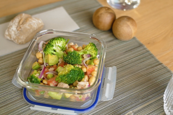 Se observa una ensalada en un tupper. La ensalada contiene garbanzos, brócoli, tomate y aguacate .