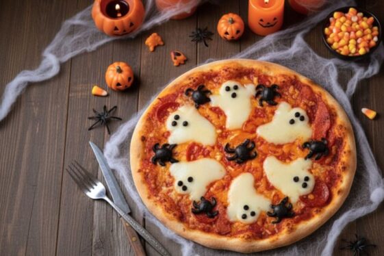 Pizza casera de Halloween decorada con fantasmas de queso mozzarella y arañas de aceituna negra, sobre base de tomate y mozzarella fundida, rodeada de decoración festiva con calabazas y velas naranjas.